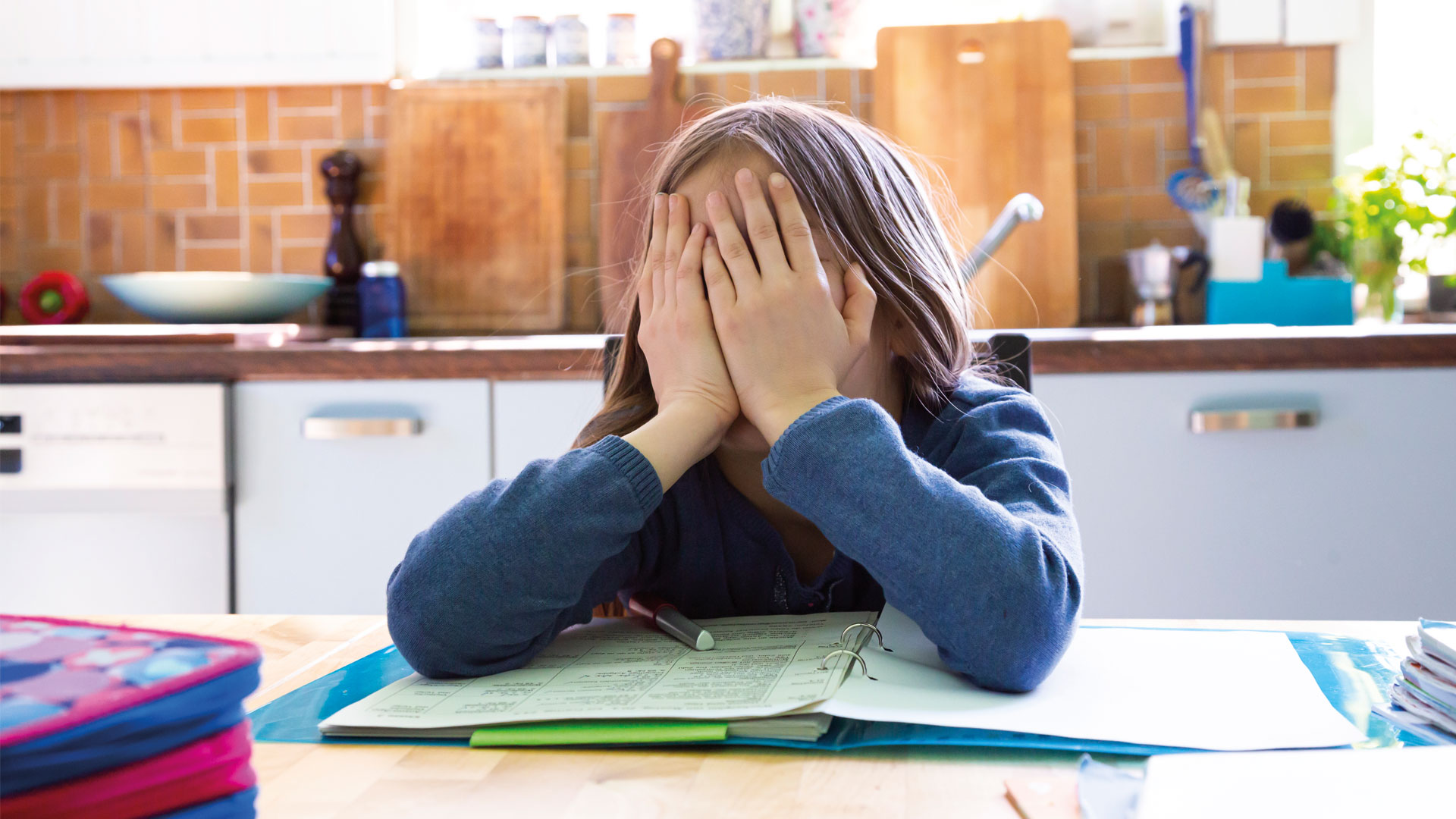 Mädchen sitzt über Schularbeiten in der Küche und hat das Gesicht in den Händen